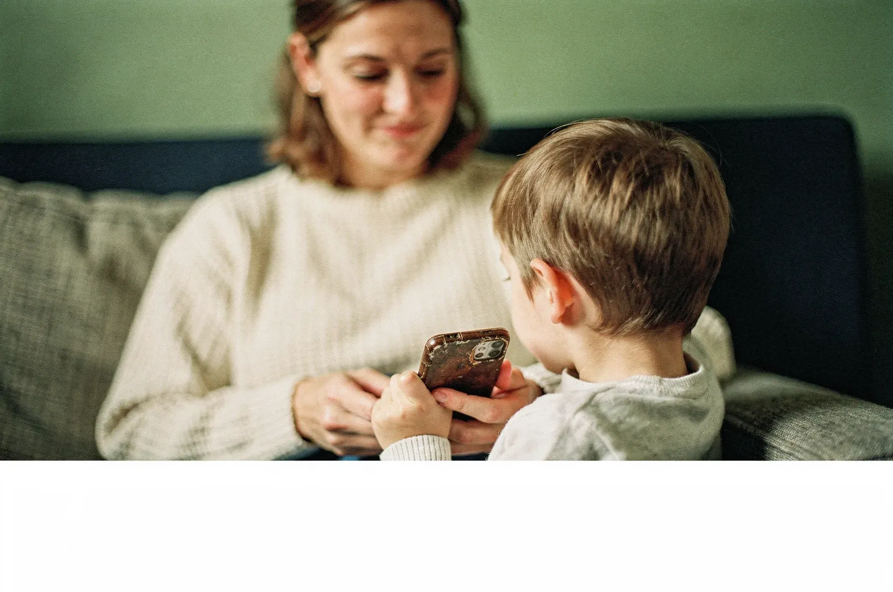 A parent and child close together in a sunlit family home, sharing an older phone.