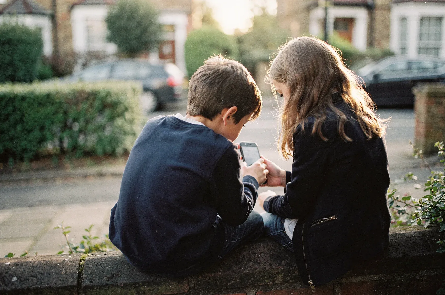Two children sit close together on a low brick garden wall, seen from behind, heads bent over a phone one of them holds, on a quiet leafy street.