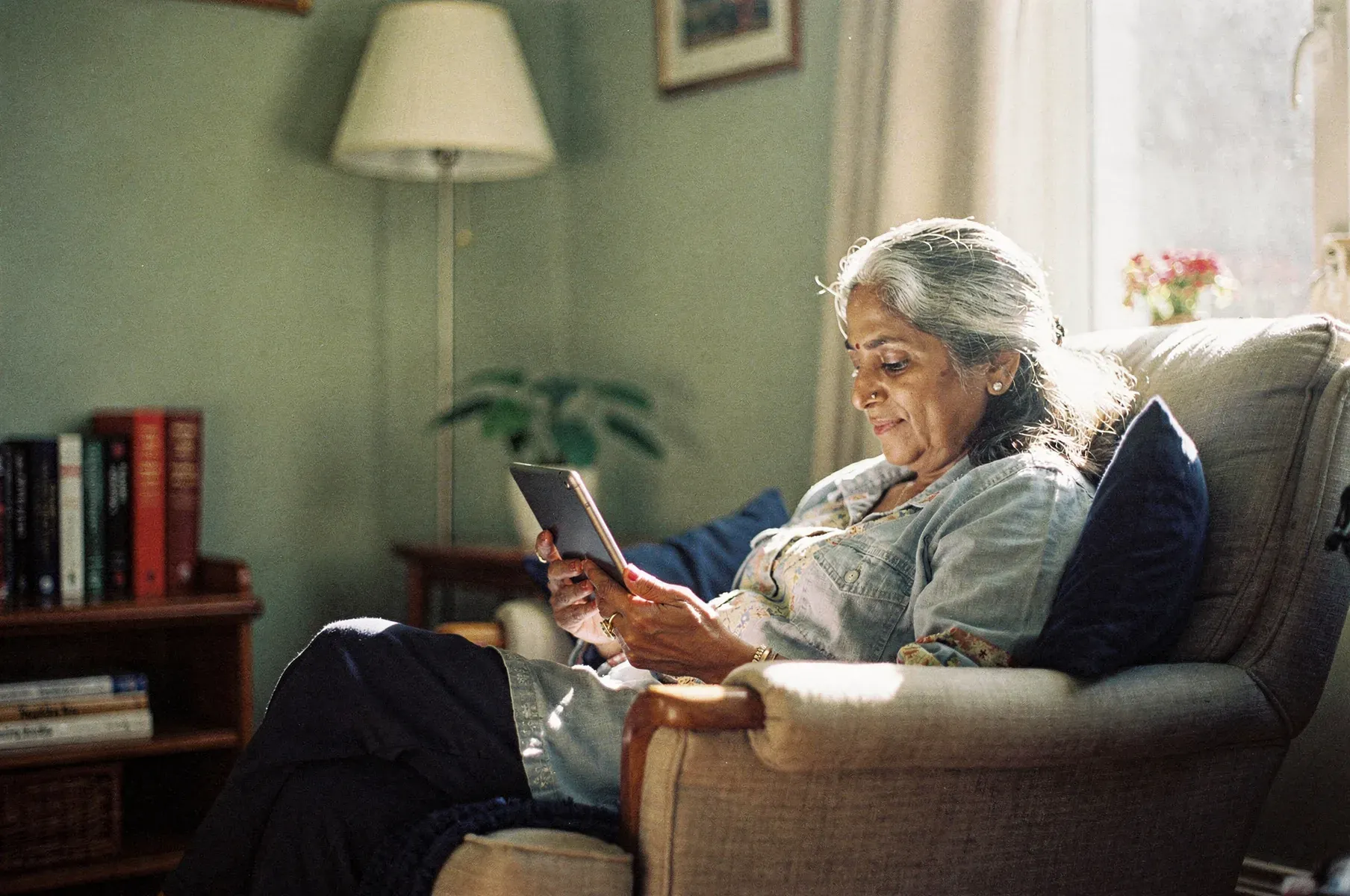 A grey-haired grandmother sits in a soft armchair in a sunlit living room, smiling at a tablet she holds, a bookcase and lamp behind her.